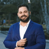 Foto de rosto de Luan sorrindo, com barba e terno escuro, em um ambiente externo